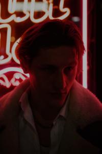 A young man, portrayed by French-British Actor Théodore Sylvain is stood in front of a red neon sign. He is blanketed in darkness, as the haze of the neon light licks the highlights and contours of his sharp features. There is a slight smirk on his face, 