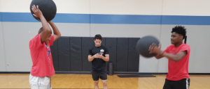 Two individuals in red shirts perform medicine ball exercises in a gym, while a third, in black, observes and records their activity.