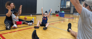 A group of children and adults are seated on a gym floor, engaging in a physical activity while using various colorful equipment scattered around.
