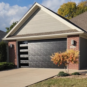Modern black garage door on a brick and gray siding house with a clean concrete driveway and small landscaped front yard.