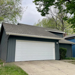 White raised-panel garage door on a dark gray house with a pitched roof, surrounded by trees and a concrete driveway.
