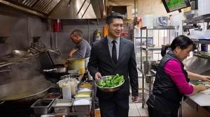 Patrick Cheng walking through a busy kitchen holding a bowl of fresh broccoli, with chefs actively preparing food in the background.