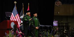 A KCU faculty member places a green and black doctoral hood on a smiling graduate during the University’s indoor commencement ceremony. U.S. and university flags are displayed on stage behind them.