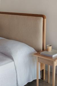 A white oak bed with an upholstered headboard in dromedary color, styles with white linen sheets, next to a square side table with a book and cup on top
