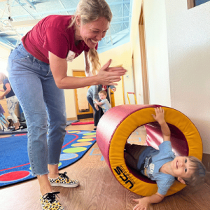A young child joyfully emerges from a colorful play tunnel, while an adult encourages them with clapping in a bright indoor play area.