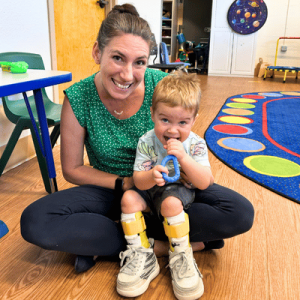 A child in a colorful T-shirt and shorts wearing braces sits on the lap of an adult, surrounded by a bright classroom setting.