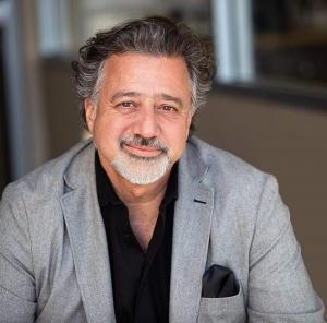 Portrait of a smiling man with salt-and-pepper hair and a short beard, wearing a light gray blazer over a black shirt with a black pocket square, seated in a softly lit indoor setting.
