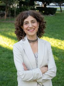 Lori Varlotta standing outdoors on a grassy area, smiling with arms crossed, wearing a light-colored pinstripe blazer, patterned blouse, and layered necklace, with trees and a pathway in the background.
