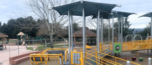 A playground featuring modern equipment, shaded structures, and nearby picnic areas, set against a cloudy sky.