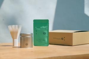 A photo of the Zenkyu Matcha subscription kit showing a paper whisk, metal tin, matcha pouch, and branded cardboard box on a wooden surface.