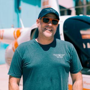 Matt Barnes, Co-Owner and Director of Education at Rogue Aviation, smiles while standing in front of a white and orange helicopter on a sunny day. He’s wearing a Rogue Aviation t-shirt, branded baseball cap, and orange sunglasses.