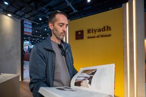 Man in casual jacket and sunglasses reads a large book in front of a yellow “Riyadh Guest of Honor” backdrop at a book fair.