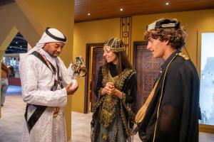 Three people in ornate Saudi attire—one pouring coffee from a brass dallah into a small cup for two others—in a fair exhibit.