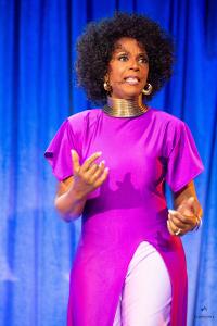 Satori Shakoor performs on stage in a vibrant purple dress and gold statement jewelry during her stand-up storytelling concert film “Confessions of a Menopausal Femme Fatale,” against a dramatic blue curtain backdrop.