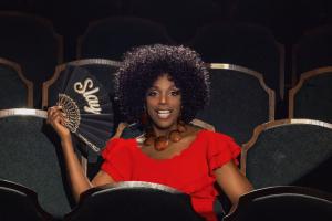 Satori Shakoor sits in a theater seat wearing a bold red dress, holding a black fan that reads “Slay.” Known for her stand-up storytelling concert film “Confessions of a Menopausal Femme Fatale,” she brings confidence and flair to the stage and screen.