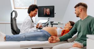 A sonographer performs an ultrasound scan on a pregnant woman, with a partner seated beside her and a 3D image visible on the monitor.