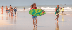A surfer carries a vibrant, spiral-patterned surfboard along a sunny beach, with others enjoying the ocean waves in the background.