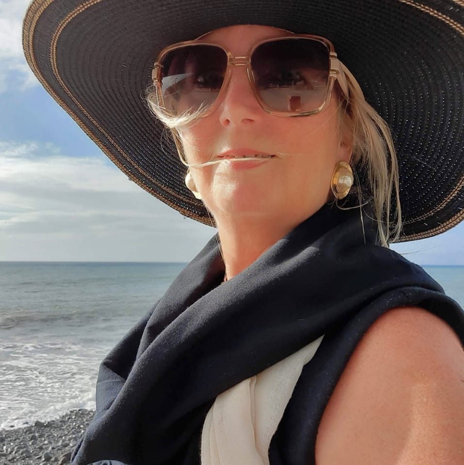 Swanette Kuntze standing at the ocean shore wearing a wide-brimmed black hat, sunglasses, and a navy scarf, gazing calmly into the distance.