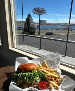 A photo of a hamburger and fries in a basket sitting on a table in The Ranch restaurant. Behind is a window looking out onto the sign for the restaurant
