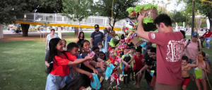 A lively gathering of children eagerly reaching for goodies from a colorful piñata in a park setting, surrounded by adults in the background.