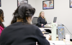 Blonde female clinician wearing a black jacket sits at a conference table with an open laptop, addressing colleagues in the foreground; Bierman Autism Centers posters hang on the wall behind her.