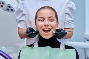 A smiling teenage girl shows her beautiful teeth after professional cleaning, with the dentist’s hands holding tools near her cheeks.