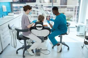 A Black male dentist explains the procedure with a teeth model as his lady assistant prepares the patient.