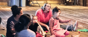 A group of children sit on a wooden floor, engaged in a creative activity while surrounded by colorful craft materials.