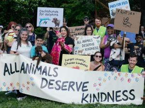 People and their pets gathered in the park behind a sign that reads "All Animals Deserve Kindness". The crowd holds various signs demanding kindness for all animals.