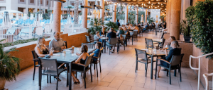Outdoor dining area bustling with families and friends, surrounded by greenery and warm lights, near a poolside setting.