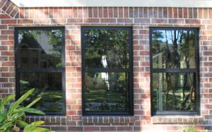 Three black-framed windows on a red brick wall reflect trees and a house under a clear sky.