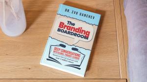 A hardcover copy of Dr. Ivo Ganchev’s book, The Branding Boardroom, placed on a wooden table next to a white vase, showcasing the cover design and subtitle: “Deep Conversations with Global Thought Leaders.