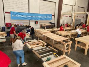Large workshop interior showing multiple volunteers wearing red shirts working at wooden tables using power tools to craft raw lumber into bed frames. A blue banner reading "NO KID SLEEPS ON THE FLOOR IN OUR TOWN!" hangs prominently across the back wall. 