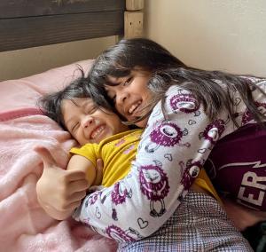 Two children with dark hair lying together on a wooden bed frame with pink bedding, both smiling broadly at the camera. One child wears a yellow shirt while the other has on pajamas with a purple and white pattern.