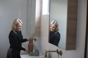 Kellie Richardson arranging a vase on a bathroom counter, smiling at her reflection in the mirror with brass tapware and minimalist décor in the background.