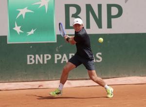 Tennis player Noah Johnston at Roland Garros 2025, focused his backhand in competition. He is blonde with a white baseball cap, wearing dark colored tennis clothes.
