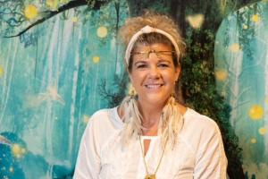 Tracey Witten, featured author in the international best-selling anthology The Call Within: Finding Purpose and Sparking Transformation, poses against a magical forest backdrop, radiating warmth and wisdom. Dressed in flowing white with feather earrings,