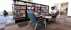 Two individuals sit in a library, engaged in conversation, surrounded by tall bookshelves filled with colorful books.
