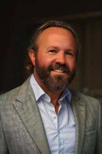 John Lufburrow, a smiling man with a short beard and medium-length hair, wearing a light gray blazer over a light blue button-down shirt.