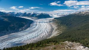 Photo shows an aerial view of the Klinaklini glacier on the central coast of British Columbia. Ice from the glacier runs down the centre of the photo in a wide valley. Snow-capped mountains and blue sky with clouds in background.