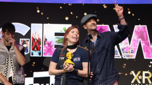 Deirdre V. Lyons and Stephen Butchko of Ferryman Collective on stage smiling and holding the Best Indie Creator trophy at the 2025 Auggie Awards during AWE, with celebration graphics behind them.