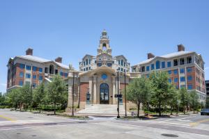 Exterior view of the Old Parkland East Campus in Dallas, Texas, featuring a grand neoclassical building with a central dome, symmetrical wings, and red brick and stone façade, framed by trees and street lamps under a clear blue sky.