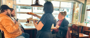 A server delivers a plate of food to seated customers in a cozy restaurant, surrounded by large windows and casual decor.