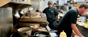 Two cooks work in a bustling kitchen, grilling meat and preparing food, with pots and pans on the counter and plates stacked in the background.