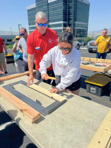 A woman wearing a white sweatshirt and sunglasses uses a hammer on a piece of lumber, guided by a man in a red SHP polo shirt and blue sunglasses. They are working at an outdoor training workshop in a parking lot with a modern glass office building in the