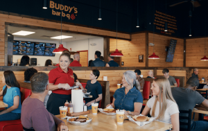 A woman standing inside a bar-b-q restaurant helping serve a table of guests