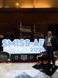 Dr. Mohana Rao Patibandla standing confidently in a suit and conference badge, positioned in front of the illuminated SMISS-AP 2025 sign featuring Mumbai landmarks like the Gateway of India, held at The Taj Mahal Palace, Mumbai, from July 10-13, 2025, wit
