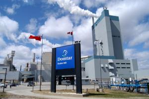 The image shows a Dominar factory in Dryden, Ontario, Canada, with the company logo in front of a headquarters building and pulp and paper production facilities