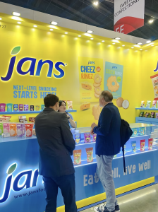 Three people converse at the Jans booth during THAIFEX 2025, with a bright yellow backdrop showcasing snack products like Cheez Ringz, Coco Ringz, and Jubes. The display emphasizes bold branding and product innovation.