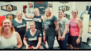 6 women and Trainer Christina Rondeau standing in a Boxing Ring